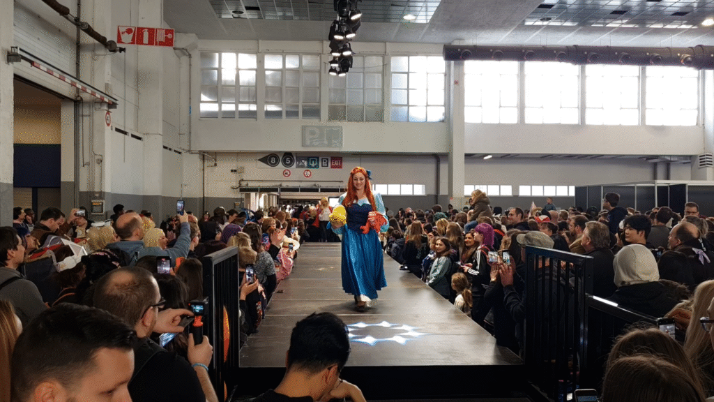 Cosplayeuse déguisée en Ariel, la Petite Sirène, défilant sur un podium avec des peluches de Polochon et Sébastien, lors d’un défilé cosplay devant un large public à la MIA à Bruxelles.